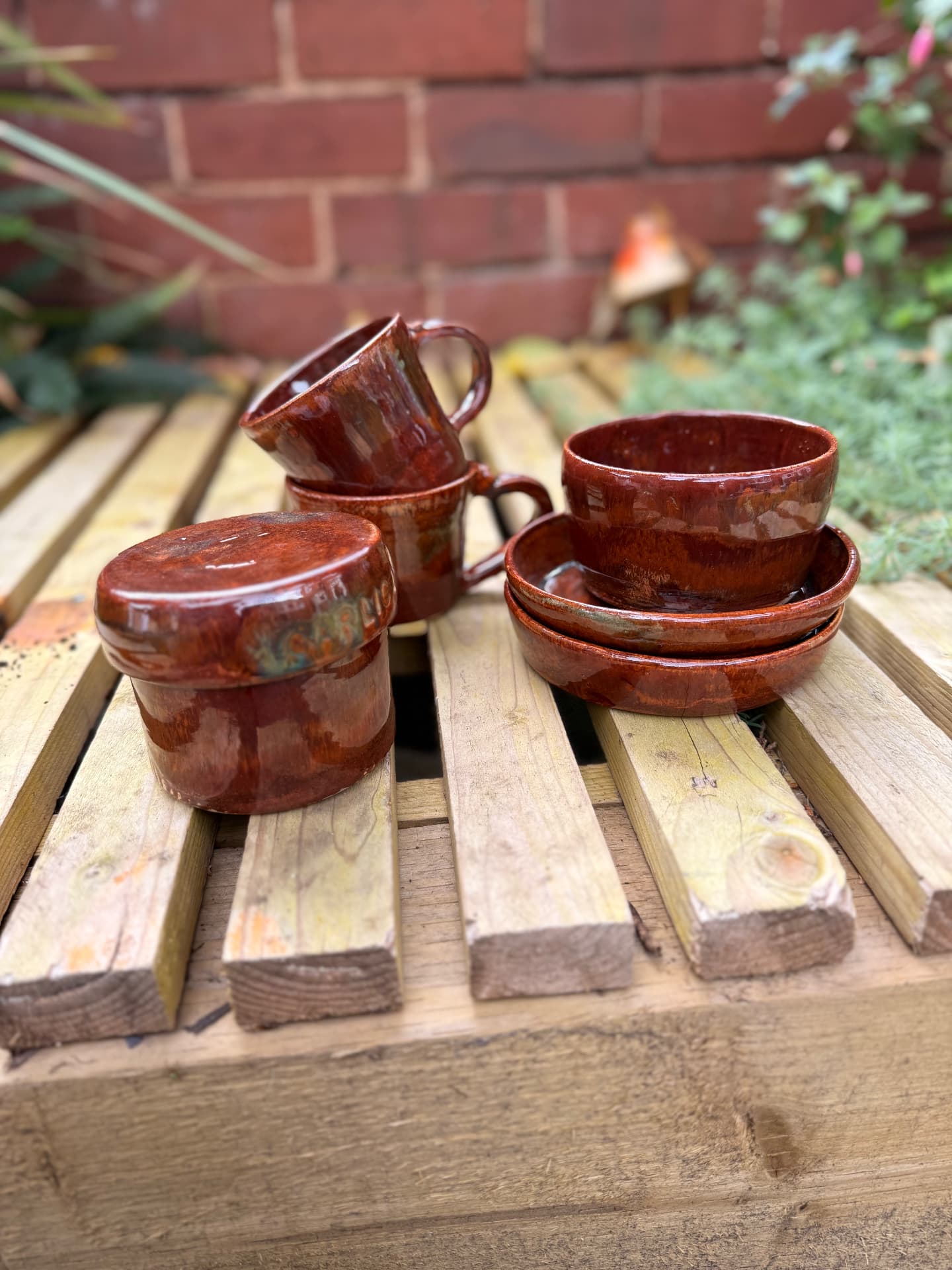 A family of rust-red pots — mugs, bowls and a lidded jar — lined up on a wooden bench